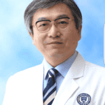 Male doctor in a white coat with glasses and a striped tie, posing in front of a blue background.