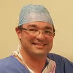 Smiling surgeon wearing scrubs and cap, ready for surgery in a hospital setting.