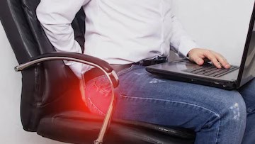 Anal Fistula 9 Person sitting on chair with lower back pain, highlighted in red, while using a laptop, indicating discomfort.